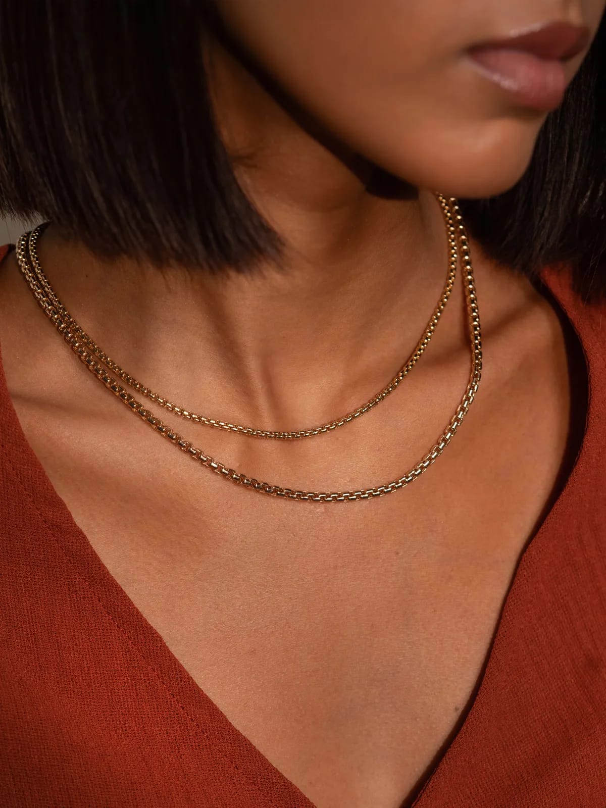 Elegant close-up shot of a woman wearing two gold chain necklaces, highlighting the sleek and sophisticated design, paired with a deep red top for a modern, stylish look.